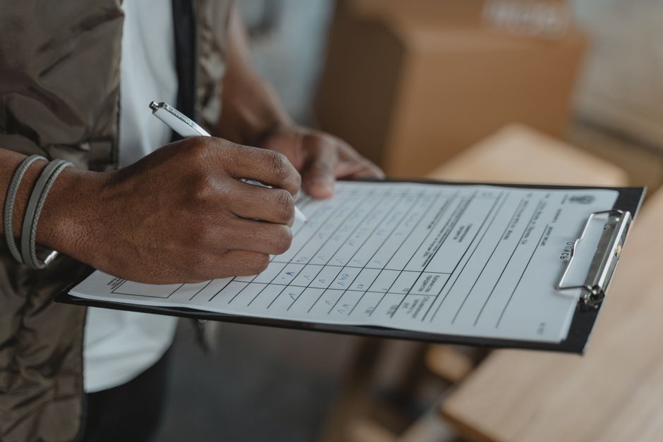 A person wearing a yellow T-shirt and jeans is in a room filled with packed cardboard boxes, some open and others closed, ready for home relocation. They are holding a clipboard in one hand and writin
